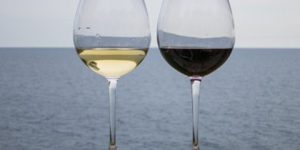 två glas vin, ett med vitt och ett med rött vin, står på ett räcke med havet i bakgrunden